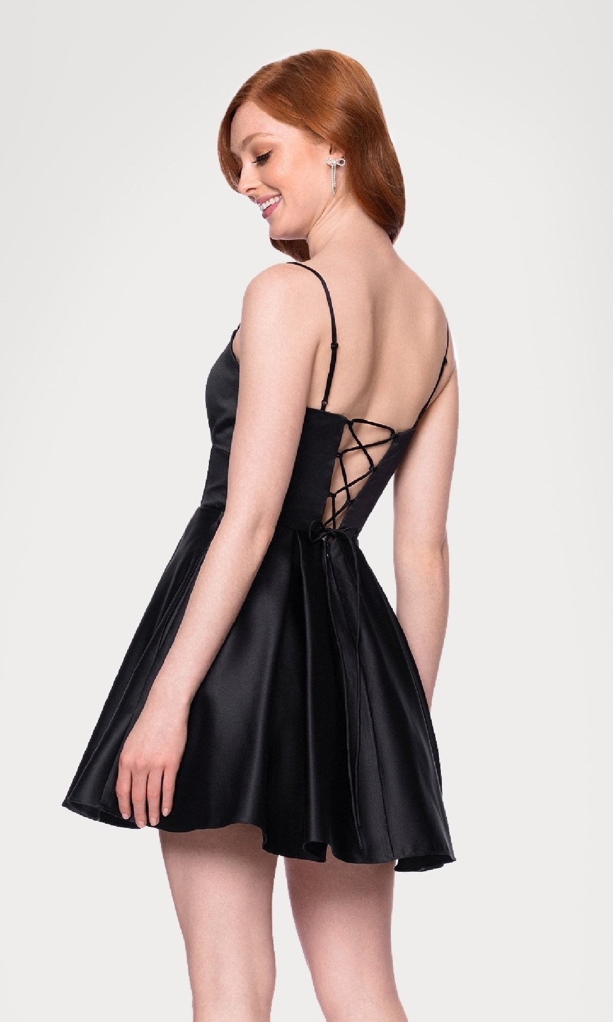 Woman wearing the Cut-Out Black Mini Hoco Dress (Blondie Nites 5605BN) with thin straps and lace-up back, standing and smiling as she looks over her shoulder.